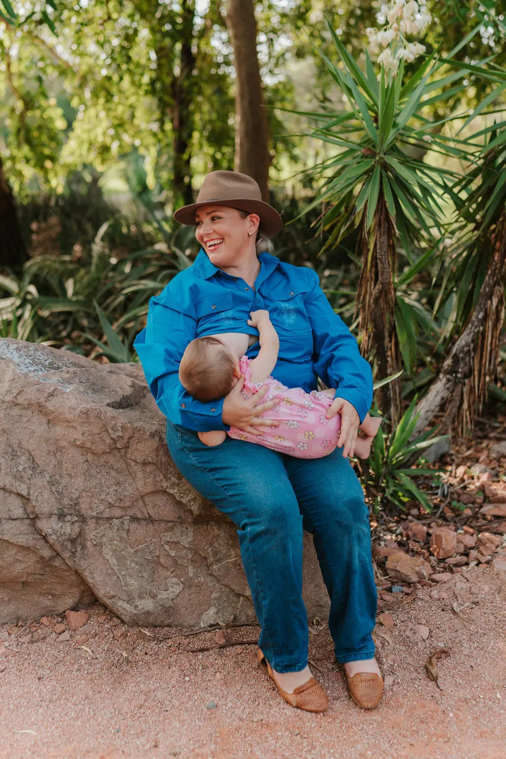 Mum nursing baby sitting on a rock outdoors in a blue long sleeve half button breastfeeding work shirt.