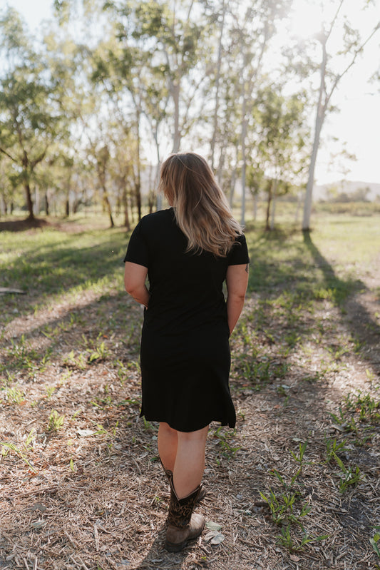 Person wearing a black dress standing in a natural setting with trees and grass.