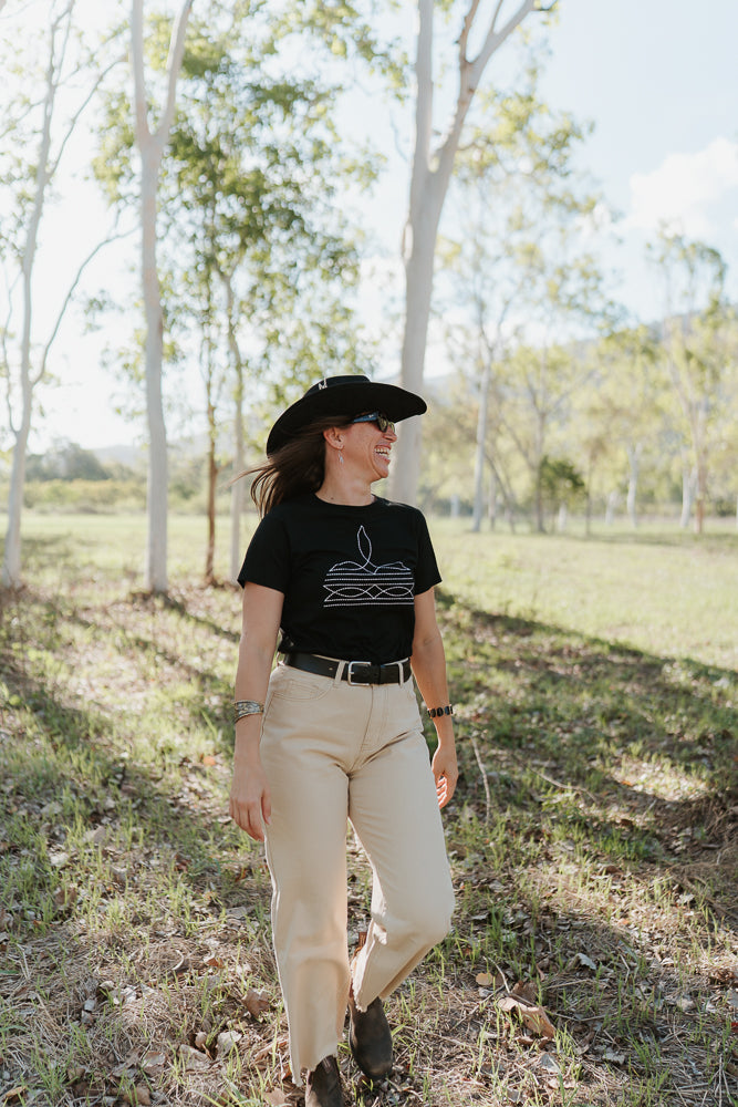 Person wearing a black t-shirt with a white design, beige pants, and a black hat in a bush setting.