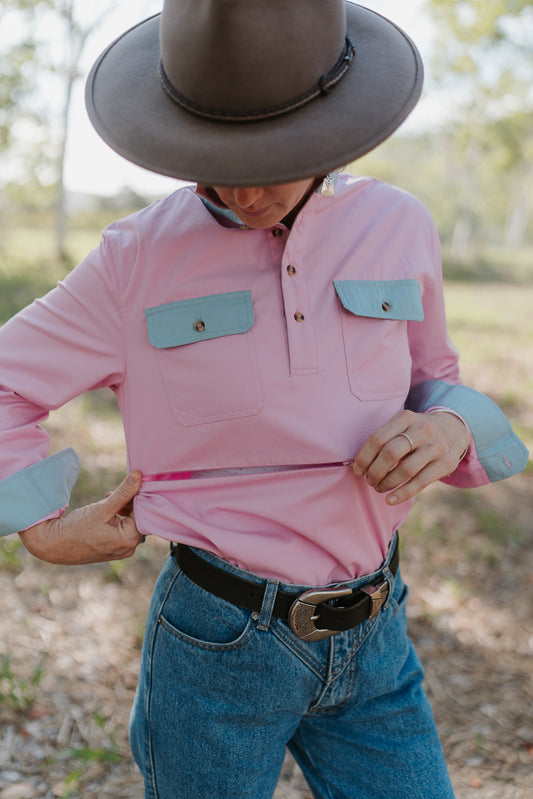 Person wearing a pink shirt with blue pocket flaps, blue jeans, and a wide-brimmed hat outdoors.