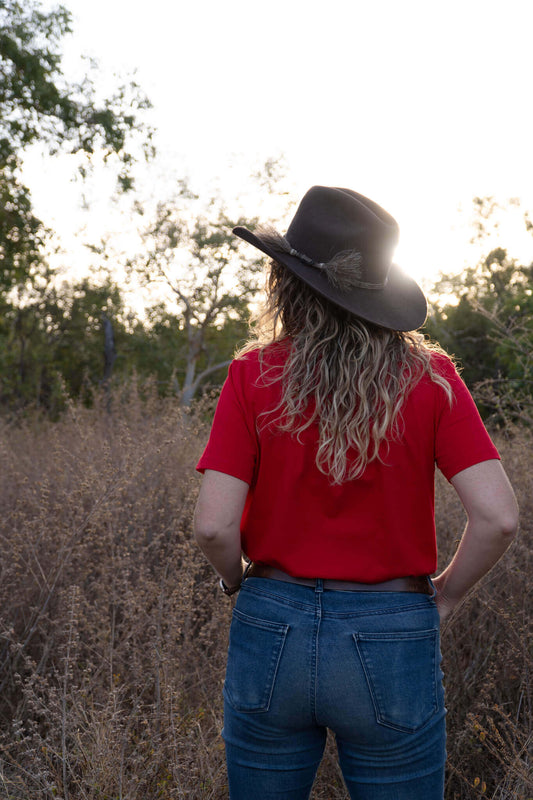 Person wearing a red shirt and blue jeans with a wide-brimmed hat, standing in a field.