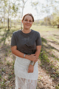 Woman standing outdoors in a natural setting wearing a dark t-shirt and light skirt.