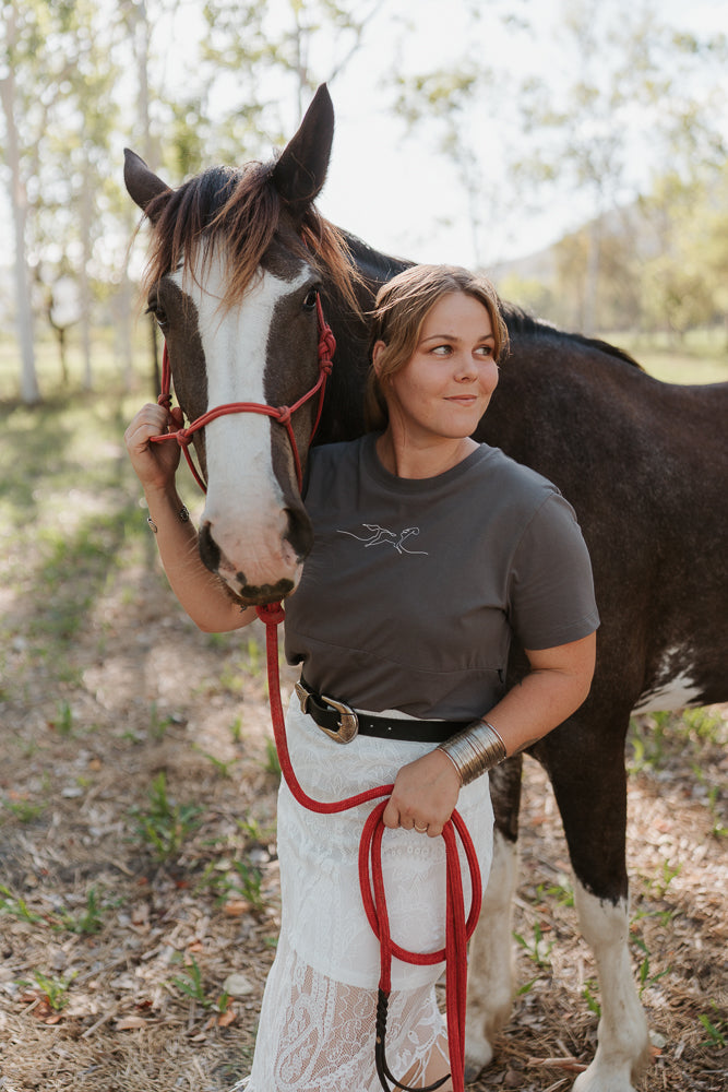 Woman standing next to a horse in an outdoor setting wearing a gray breastfeeding shirt. 