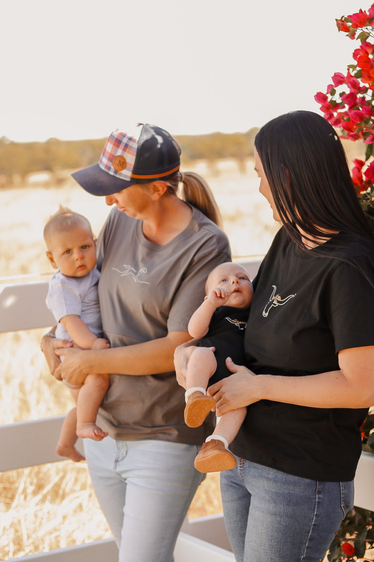 Two mums standing on a cattle property wearing mum wearing a tittytees breastfeeding shirts holding babies with matching onesies.