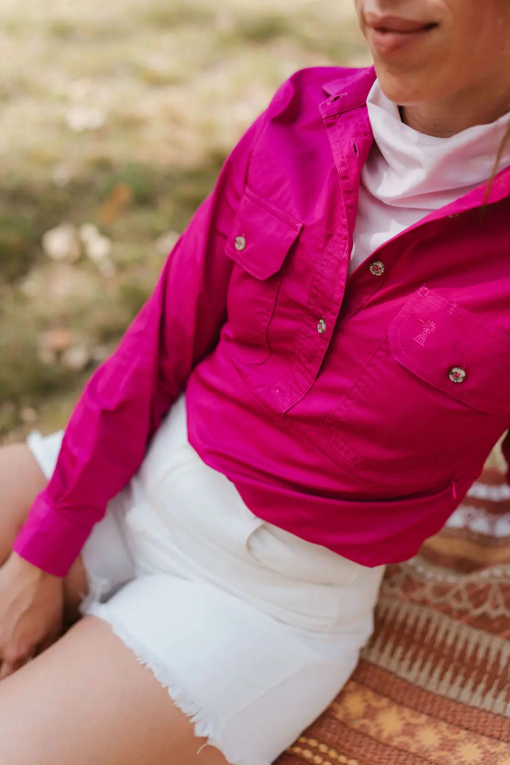 Person wearing a bright pink shirt and white shorts sitting on a patterned surface with a blurred natural background.