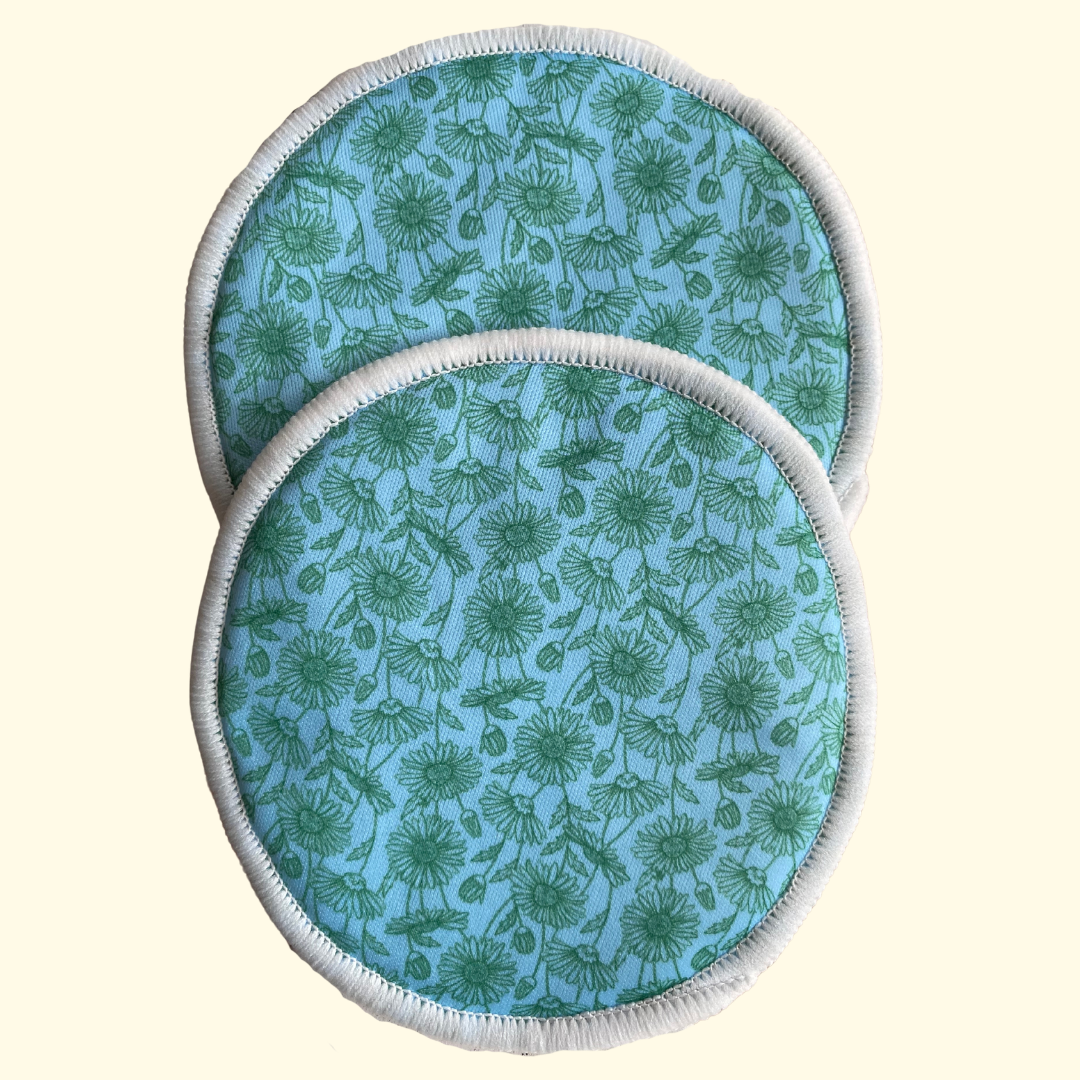 Two round reusable nursing or feeding pads with a light blue background and green dandelion pattern, finished with white stitched edges, shown on a pale cream background.
