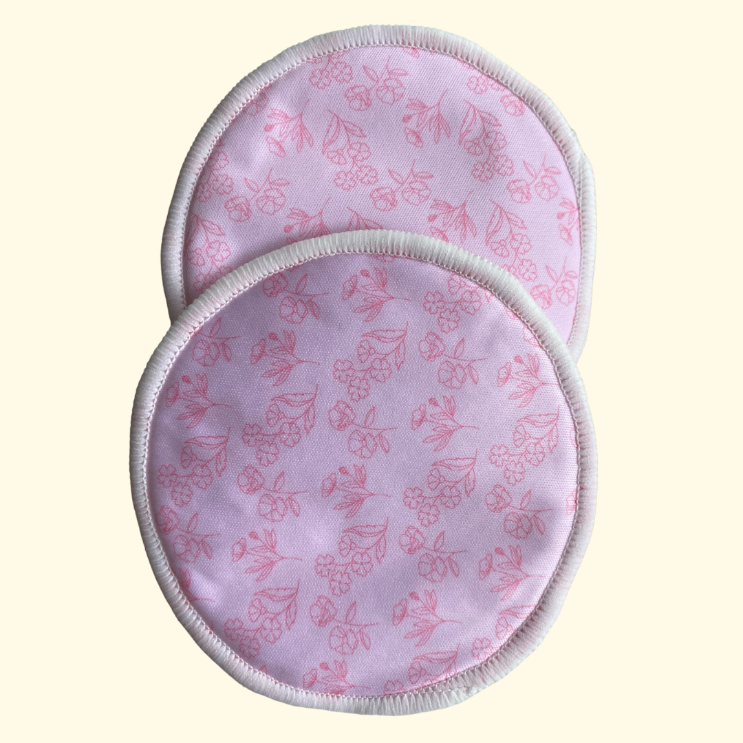 Two round reusable nursing or breastfeeding pads with a pink floral pattern on a light pink background, finished with white piped edges, shown on a pale cream background.