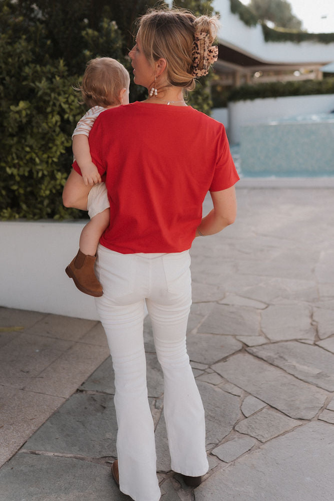 Woman in a red breastfeeding shirt and white pants carrying a child on a stone path.