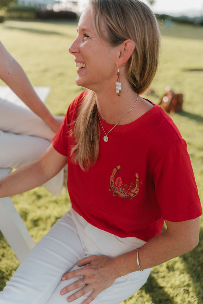 Woman wearing a red breastfeeding shirt with a design, sitting outdoors on grass.