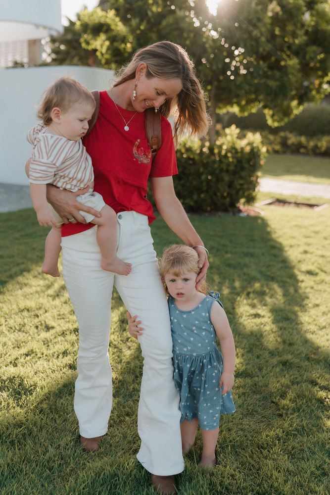 Woman in a red nursing shirt and white pants holding two children outdoors on a sunny day.