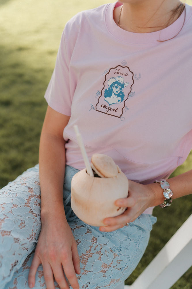 Person wearing a pink nursing t-shirt with a graphic design, holding a coconut with a straw, sitting on a white bench.