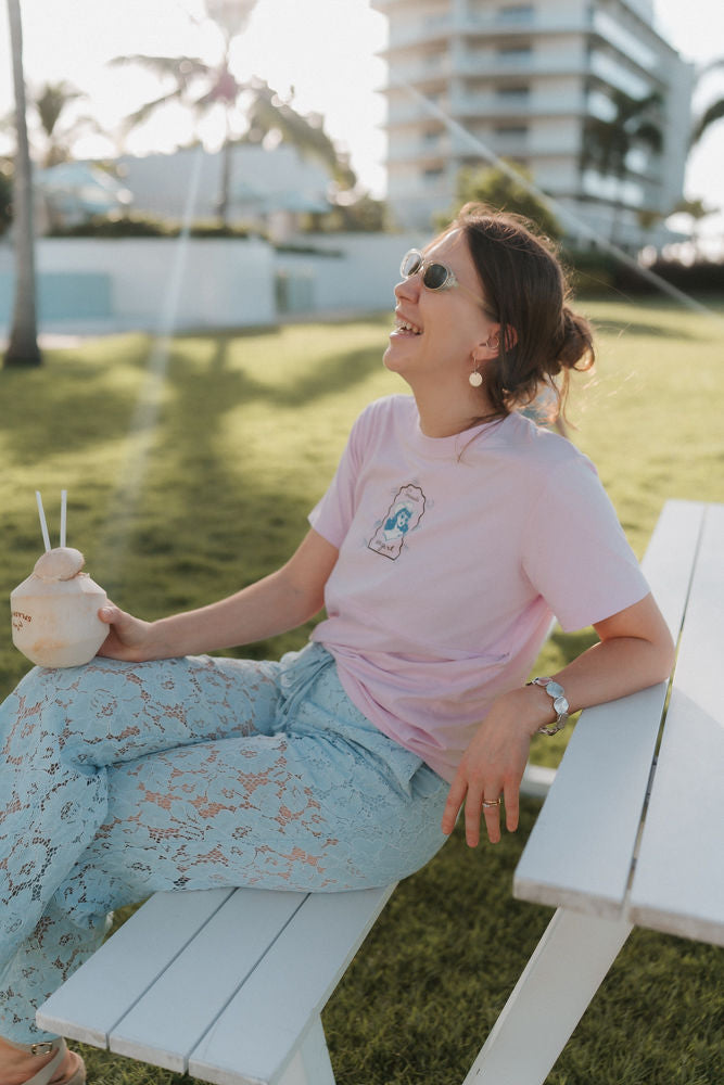 Woman sitting on a bench outdoors holding a drink, wearing a pink breastfeeding t-shirt and light blue jeans.