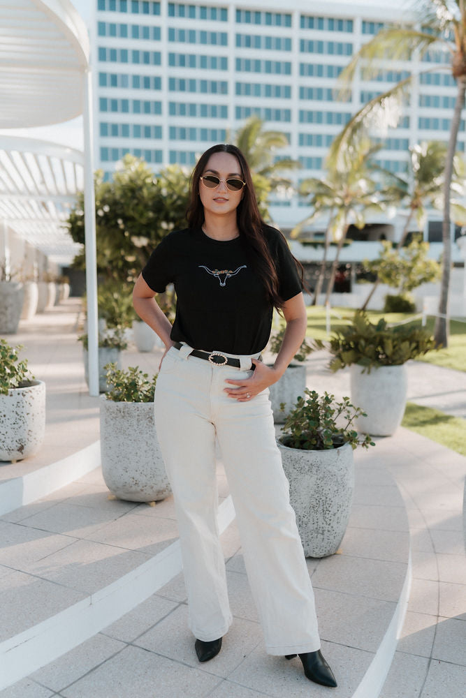 Woman in black nursing t-shirt and white pants standing on a rooftop with plants and buildings in the background