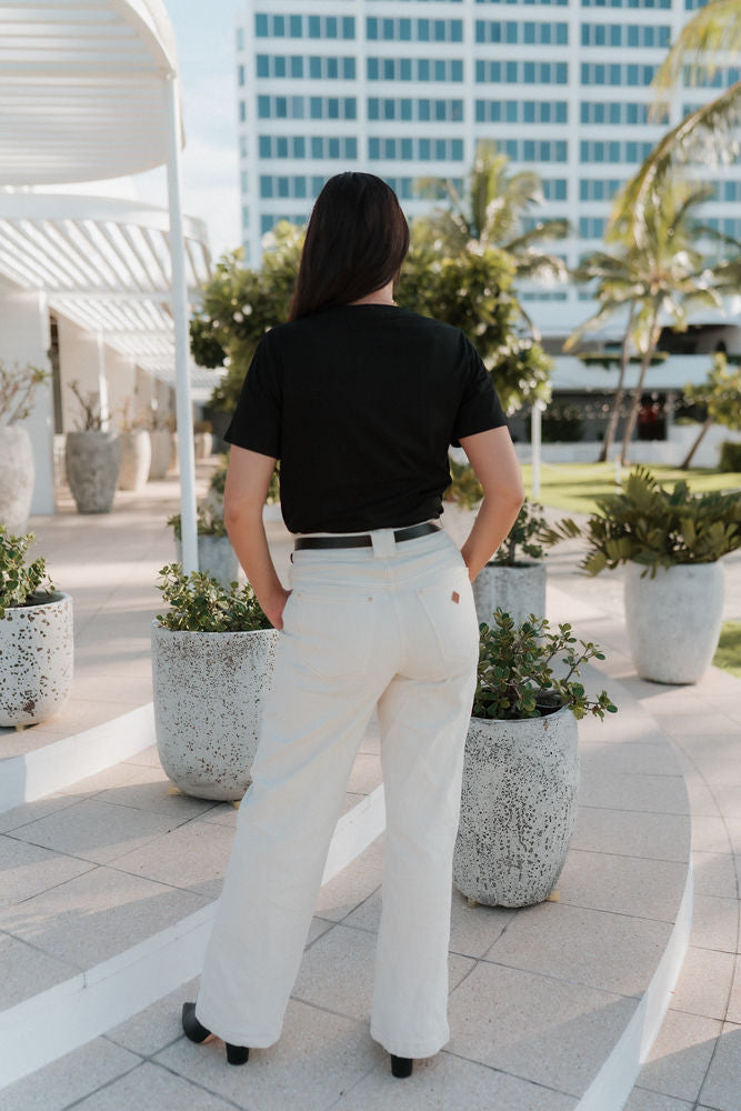 Person wearing a black shirt and white pants standing on a patio with potted plants and a building in the background.