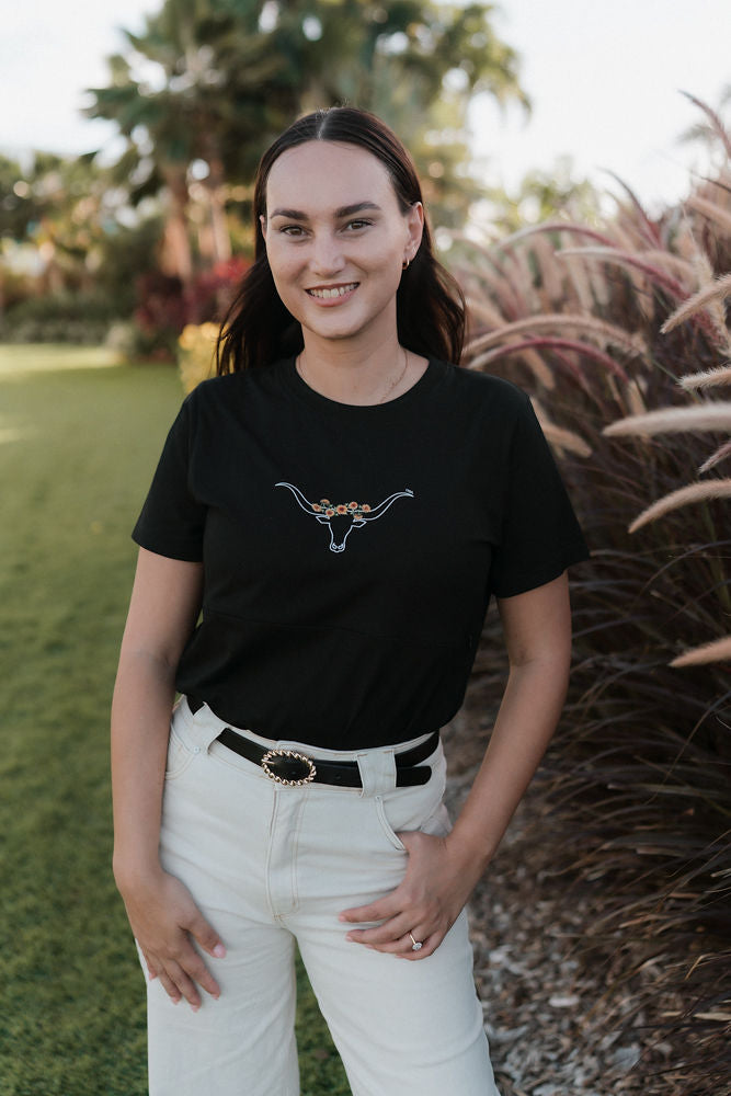 Woman wearing a black nursing t-shirt with a cowhorn annd sunflower design and white pants standing outdoors.