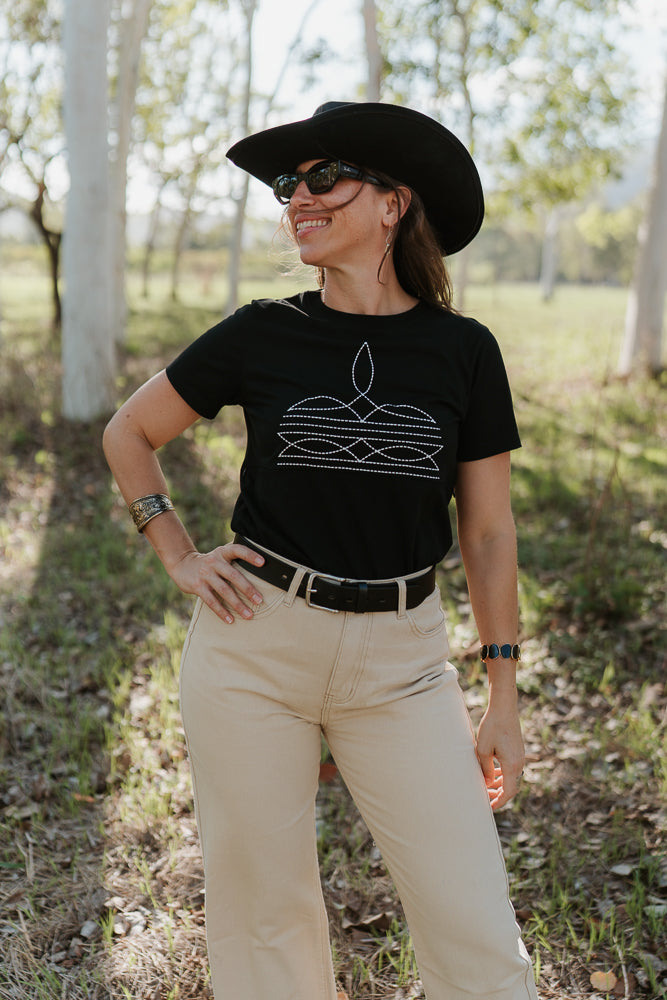 Woman wearing a black t-shirt with a graphic design, beige pants, and a black cowboy hat outdoors.