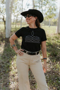 Woman wearing a black t-shirt with a graphic design, beige pants, and a black cowboy hat outdoors.