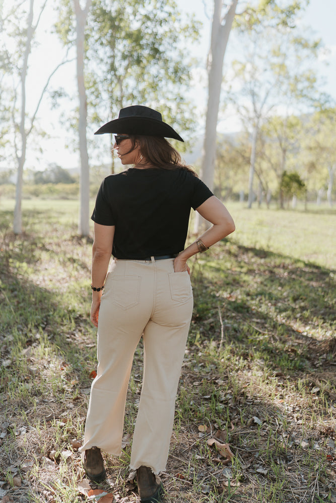 Person wearing a black cowboy hat, black shirt, and beige pants standing in a grassy field.