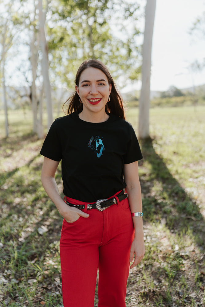 Woman wearing a black nursing t-shirt with a blue logo and red pants standing in a forest.