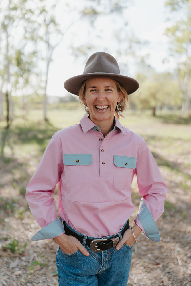 Person wearing a pink shirt with blue pocket flaps and a brown hat outdoors.