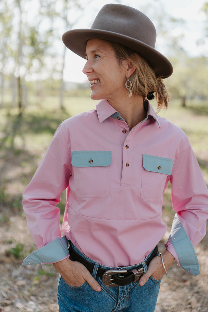 Person wearing a pink shirt with blue pocket accents and a brown hat outdoors.