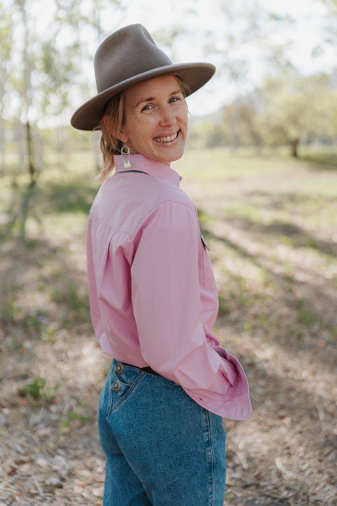 Woman wearing a pink shirt and blue jeans with a brown hat in an outdoor setting.