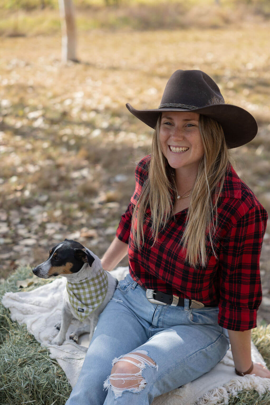 Woman in a red plaid shirt and cowboy hat sitting with a dog on a blanket in a field.