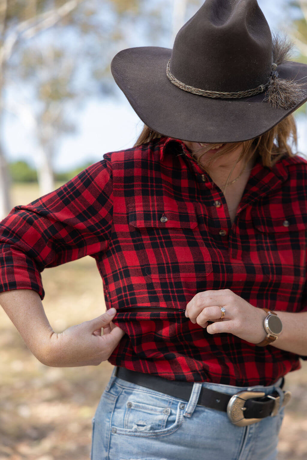 Person wearing a red plaid shirt, brown hat, and jeans outdoors. Showing how the breastfeeding zip opens.