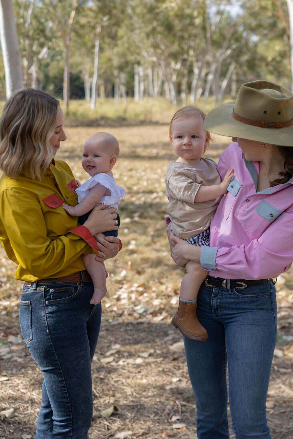 Two women holding babies outdoors with trees in the background