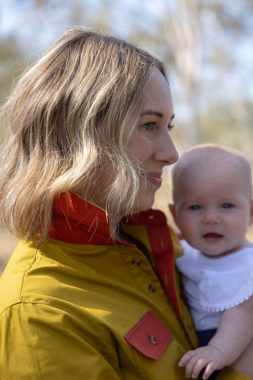 Woman in a mustard and red Breastfeeidng shirt holding a baby outdoors