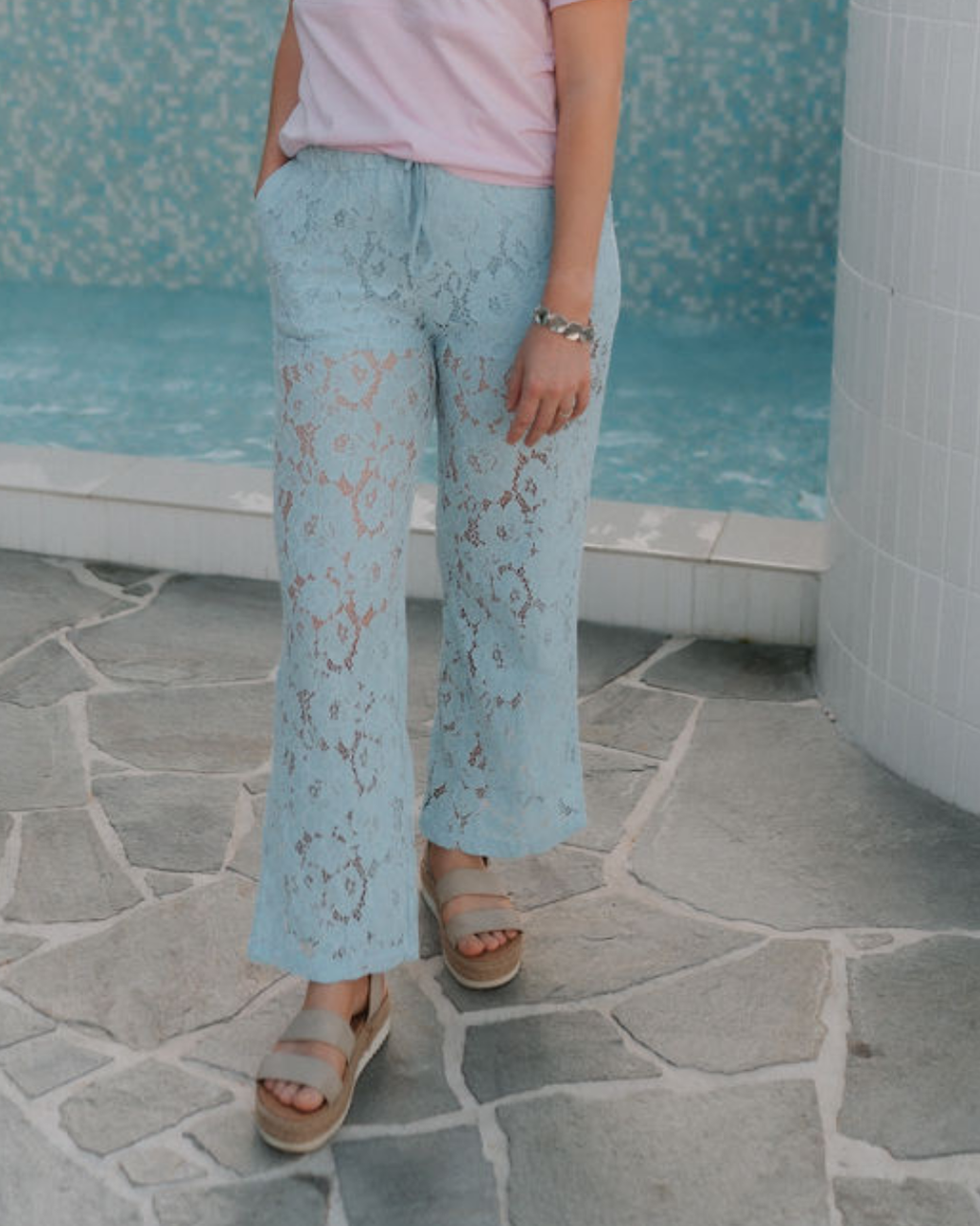Person wearing light blue lace postpartum pants and a pink top, standing on a stone floor with a pool in the background.