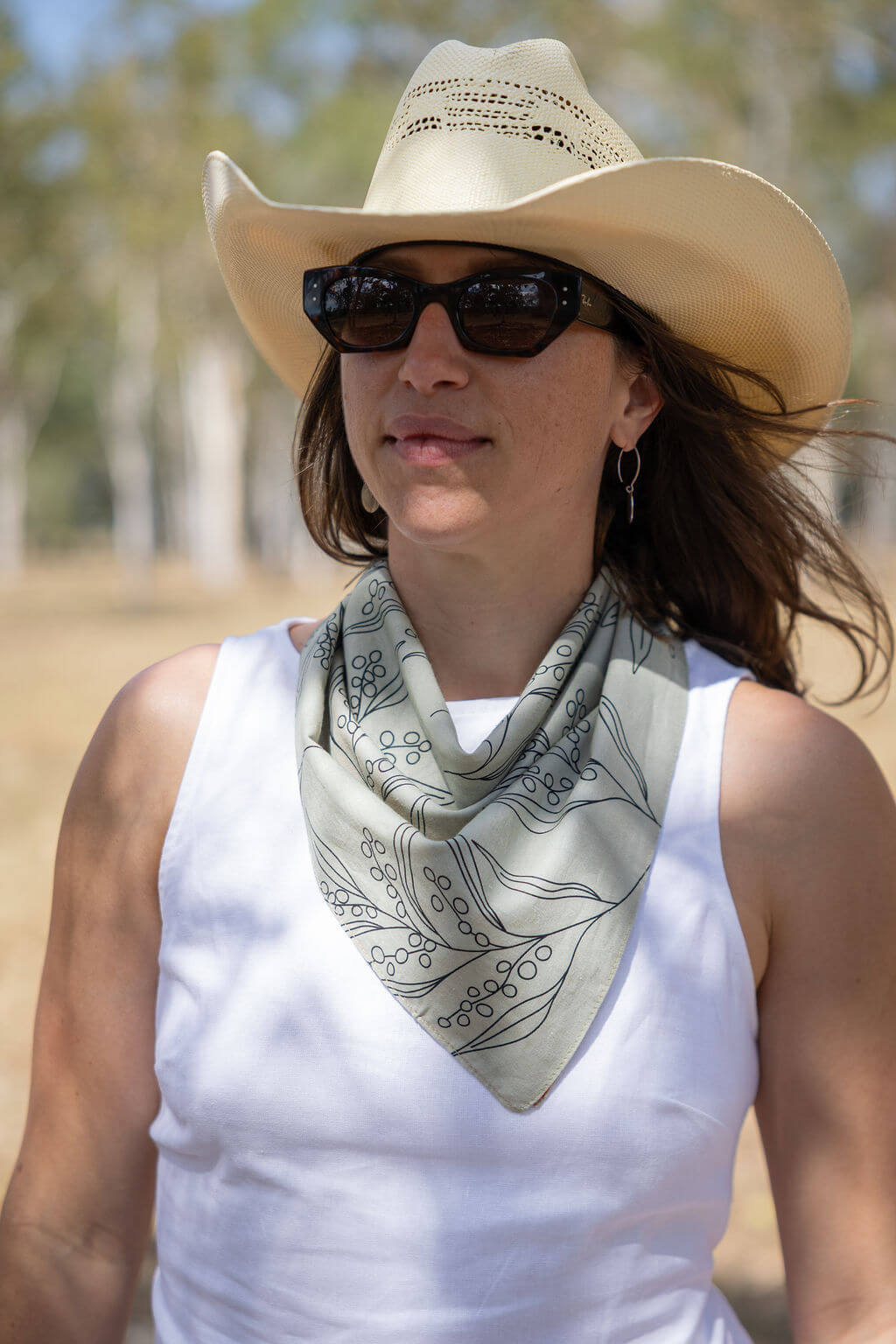 Woman wearing a cowboy hat and sunglasses with a patterned scarf, outdoors.